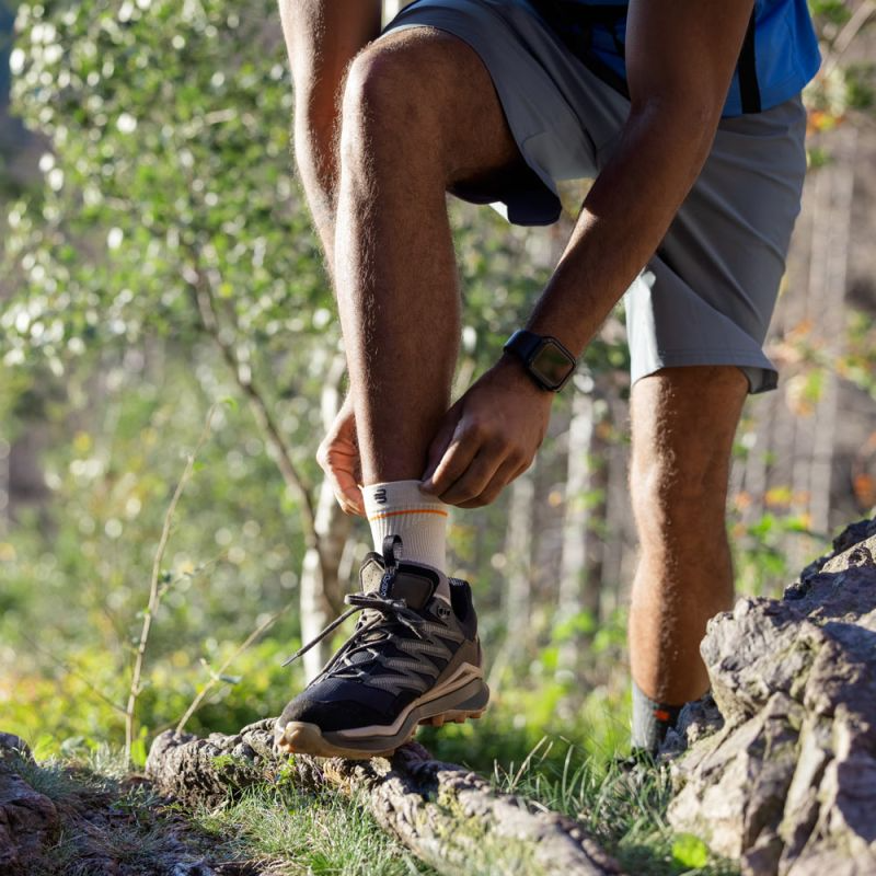 A close-up outdoor shot of a person hiking in a forest, adjusting their Bauerfeind Sport Outdoor Ankle Compression Sleeve. The person is wearing gray shorts, a blue shirt, and a black digital watch.