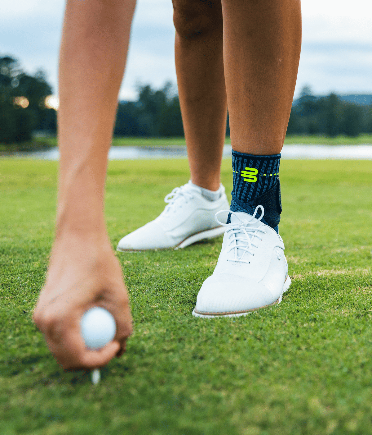 A close-up lifestyle shot of a golfer's legs on a green golf course, wearing white golf shoes and the bauerfeind sports ankle support. One hand is visible holding a golf ball on a tee.