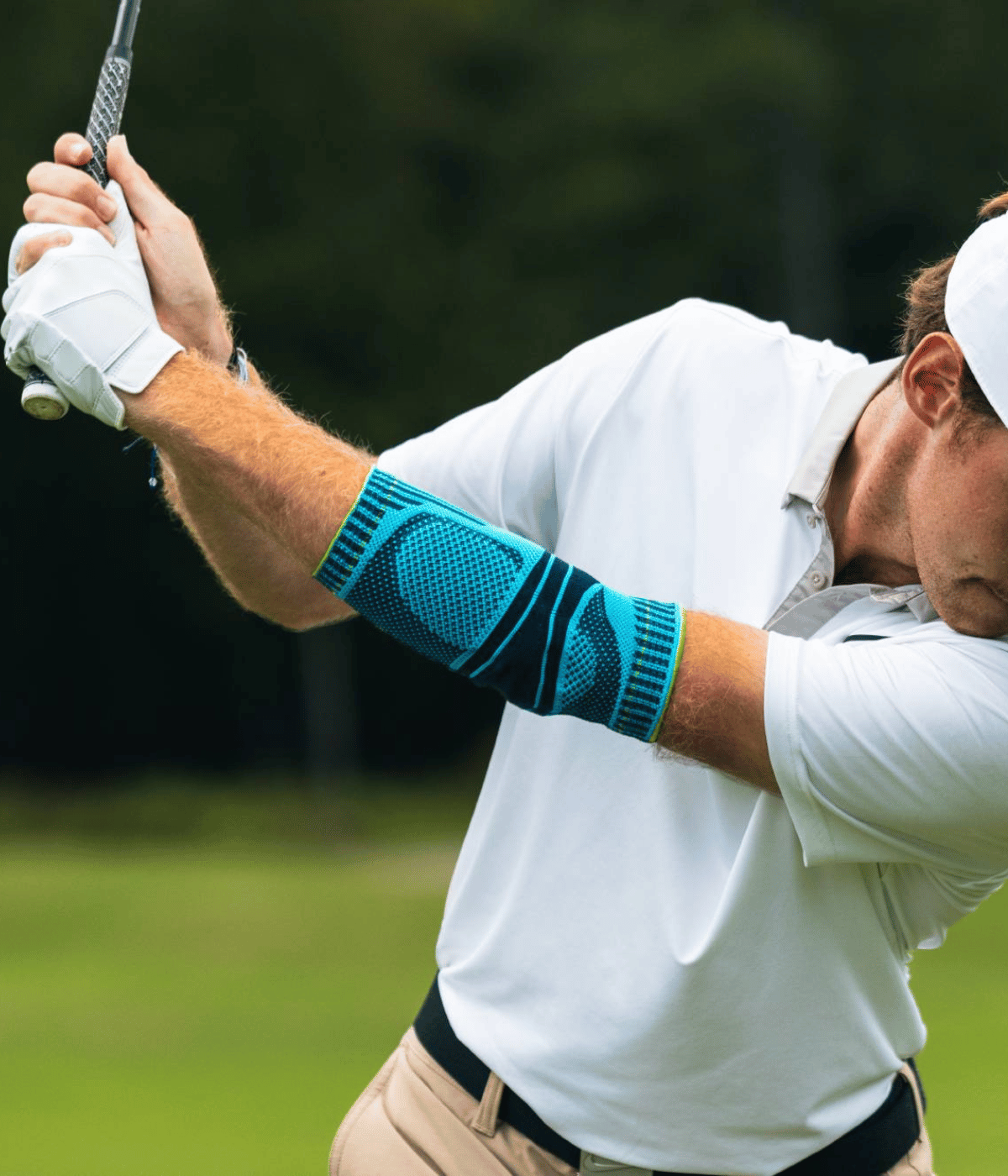 A man wearing a white polo shirt and beige pants swings a golf club on a grassy field, wearing a Bauerfeind Sports Elbow Support