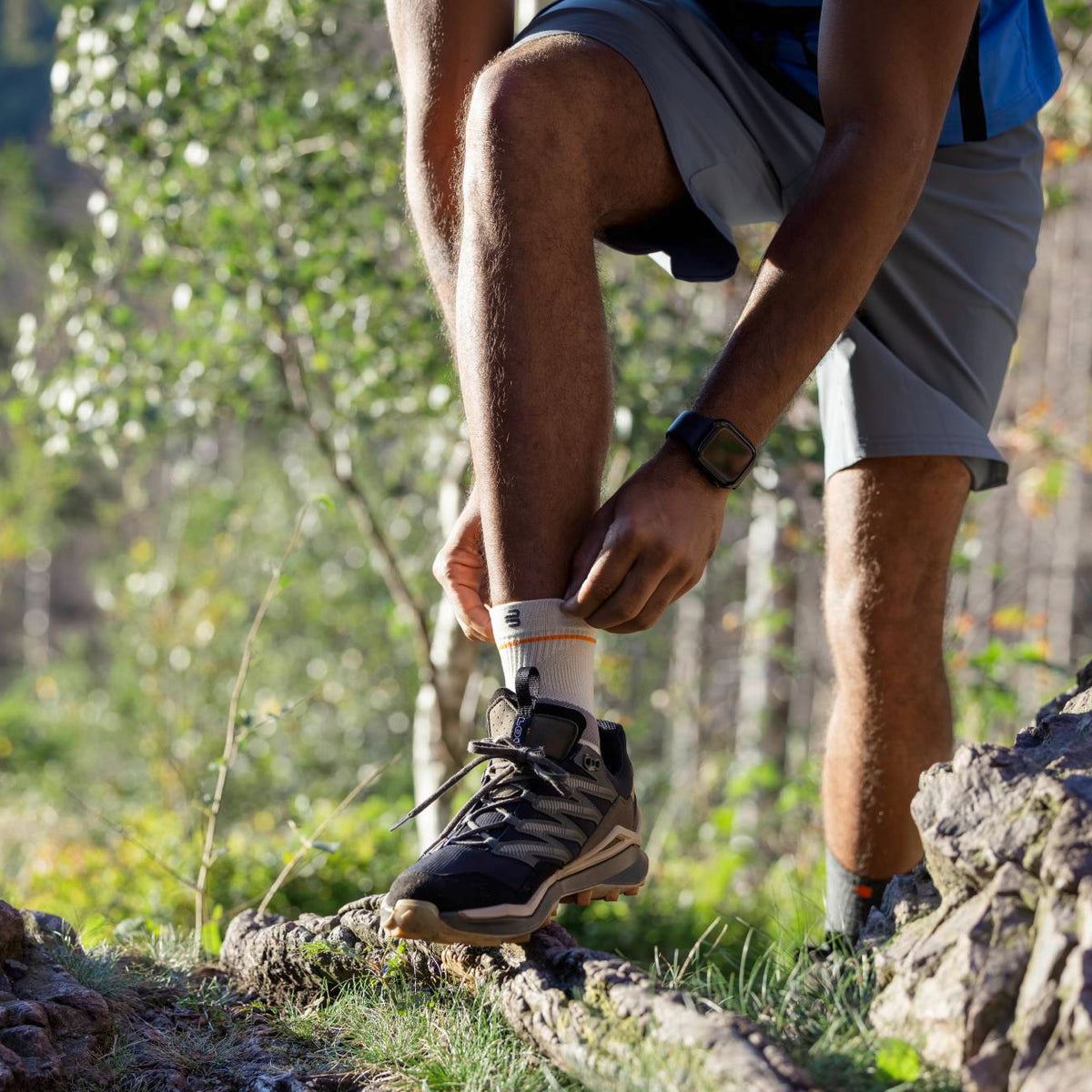 A close-up outdoor shot of a person hiking on a rocky trail, adjusting his Bauerfeind sport outdoor compression ankle sleeve. The person is wearing gray shorts and a blue shirt, with a blurred forest background.