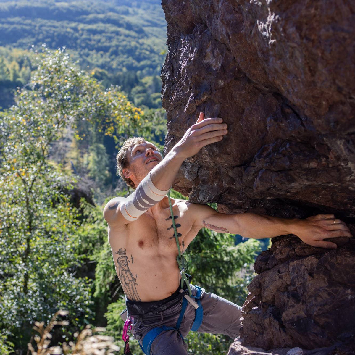 A male rock climber wearing Bauerfeind Sport Outdoor Compression Elbow Sleeve is scaling a large brown rock formation outdoors, with a lush green forested mountain valley in the background.