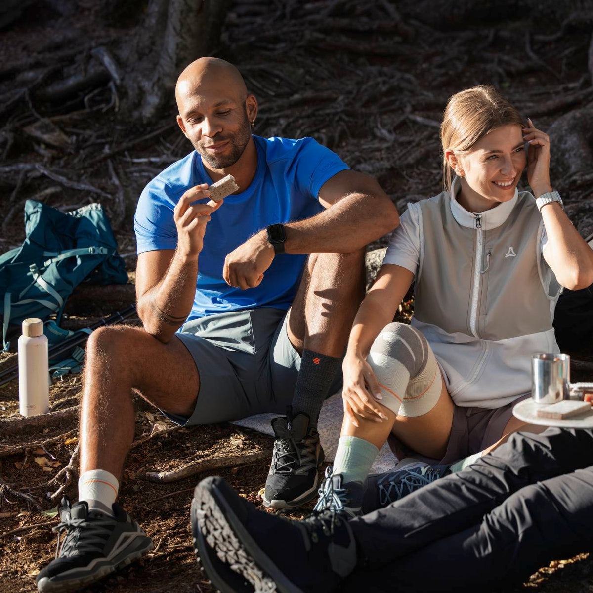Two people sitting outdoors in a wooded setting, wearing Bauerfeild sport compression ankle sleeve and knee support interacting while eating and drinking.