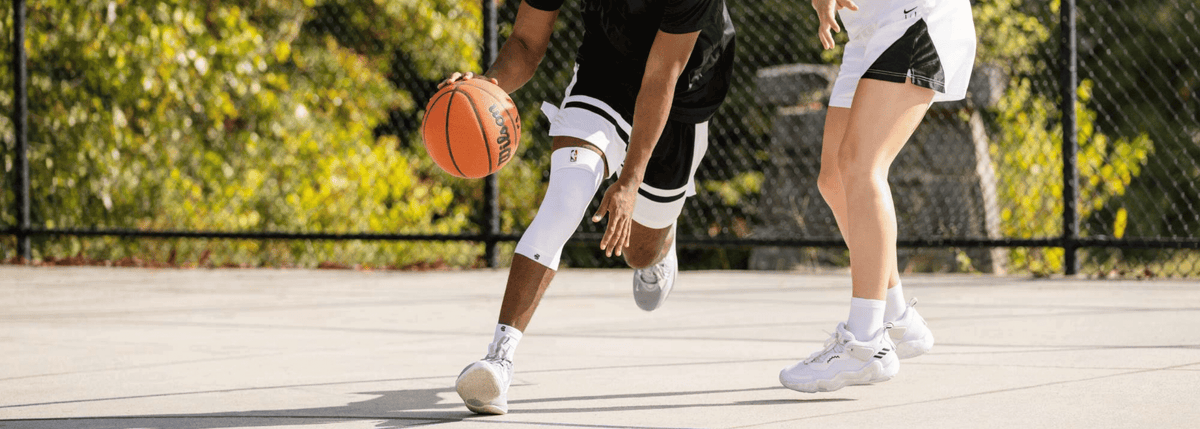 Two people playing basketball on an outdoor court with trees in the background. one of the people are wearing the White Bauerfeind Sports Compression Knee Support NBA Sleeve