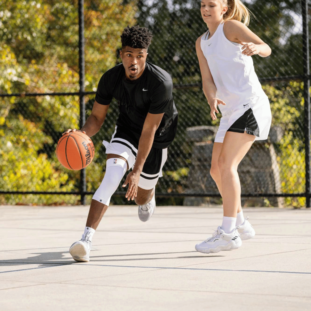 Two people playing basketball on an outdoor court with trees in the background. one of the people are wearing the White Bauerfeind Sports Compression Knee Support NBA Sleeve
