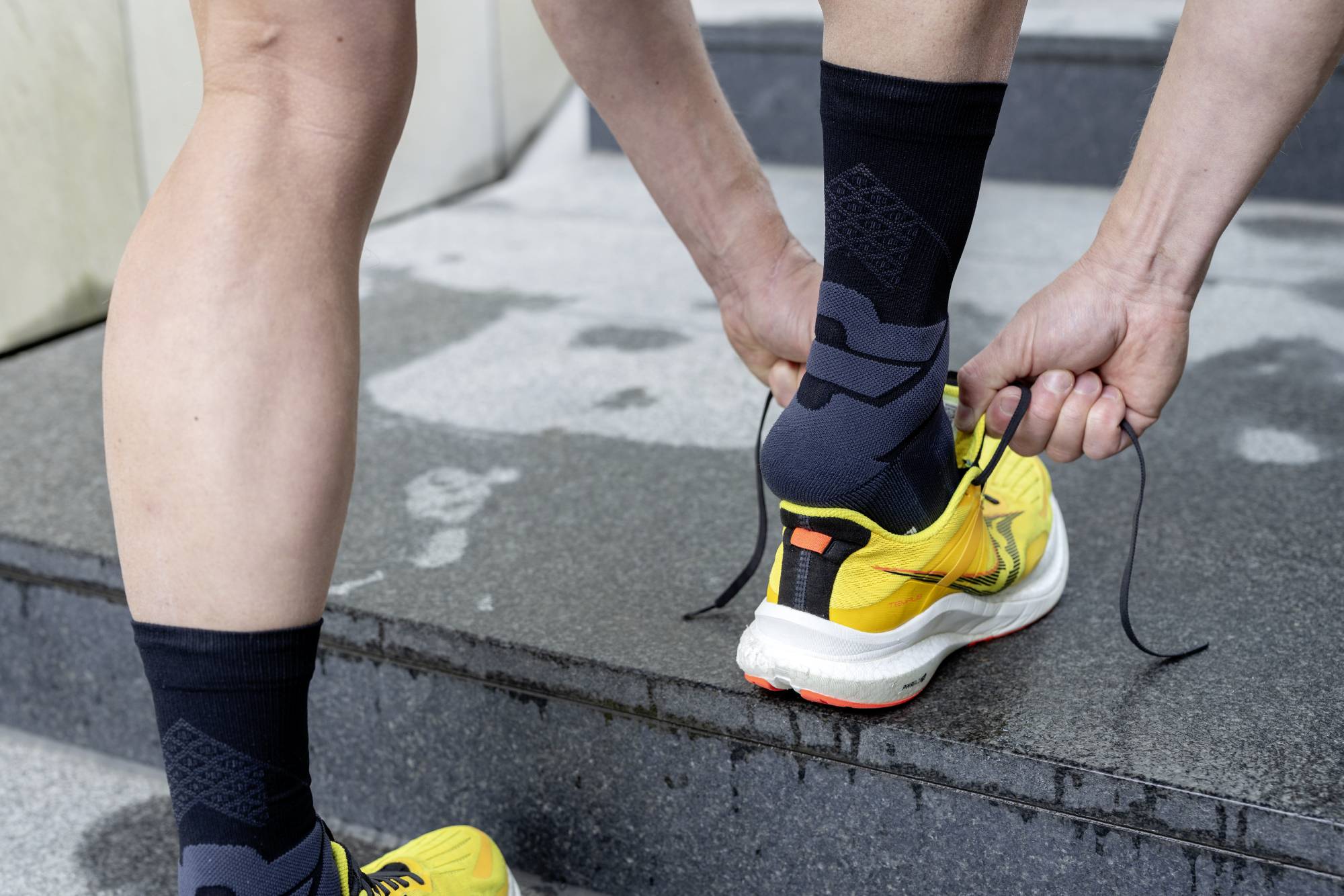 Person wearing Black bauerfeind mid-cut run performance compression socks and yellow running shoes on a concrete surface
