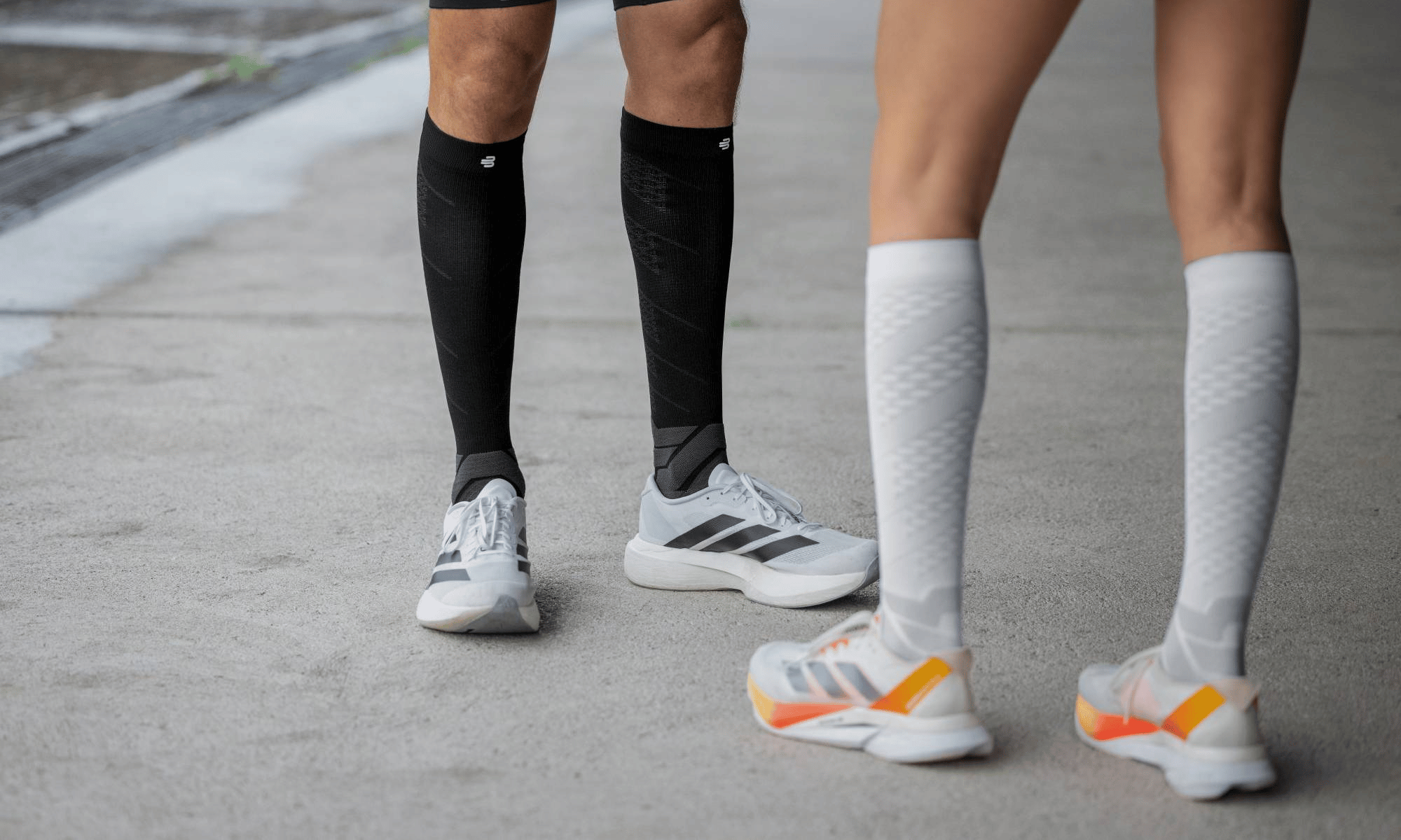 Two pairs of legs wearing bauerfeind Run Performance compression socks and athletic shoes on a pavement.