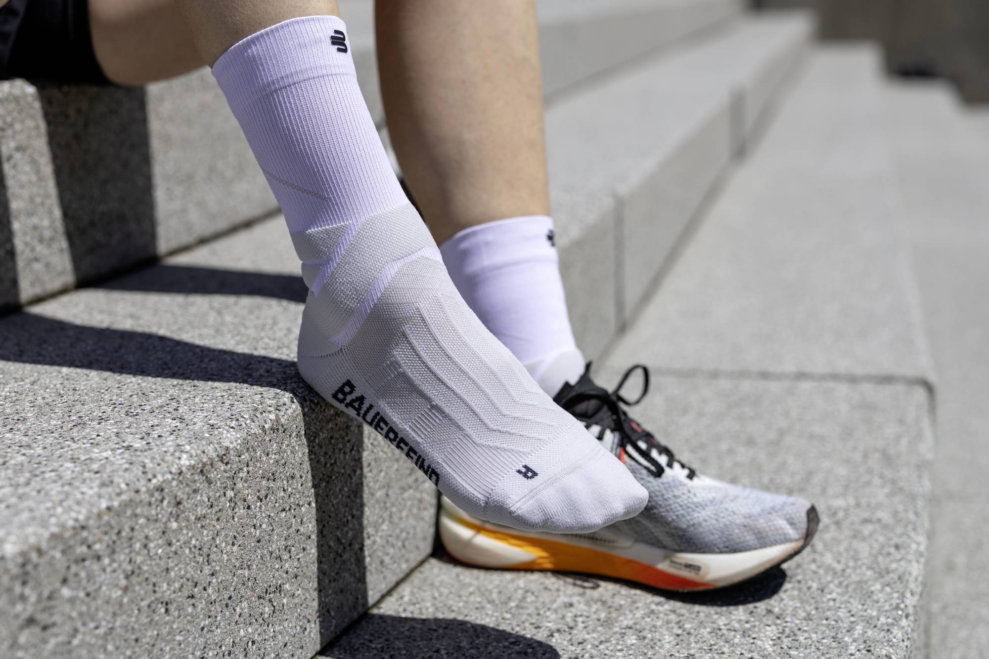 Person wearing white bauerfeind mid-cut run performance compression socks and gray sneakers with orange sole on concrete steps