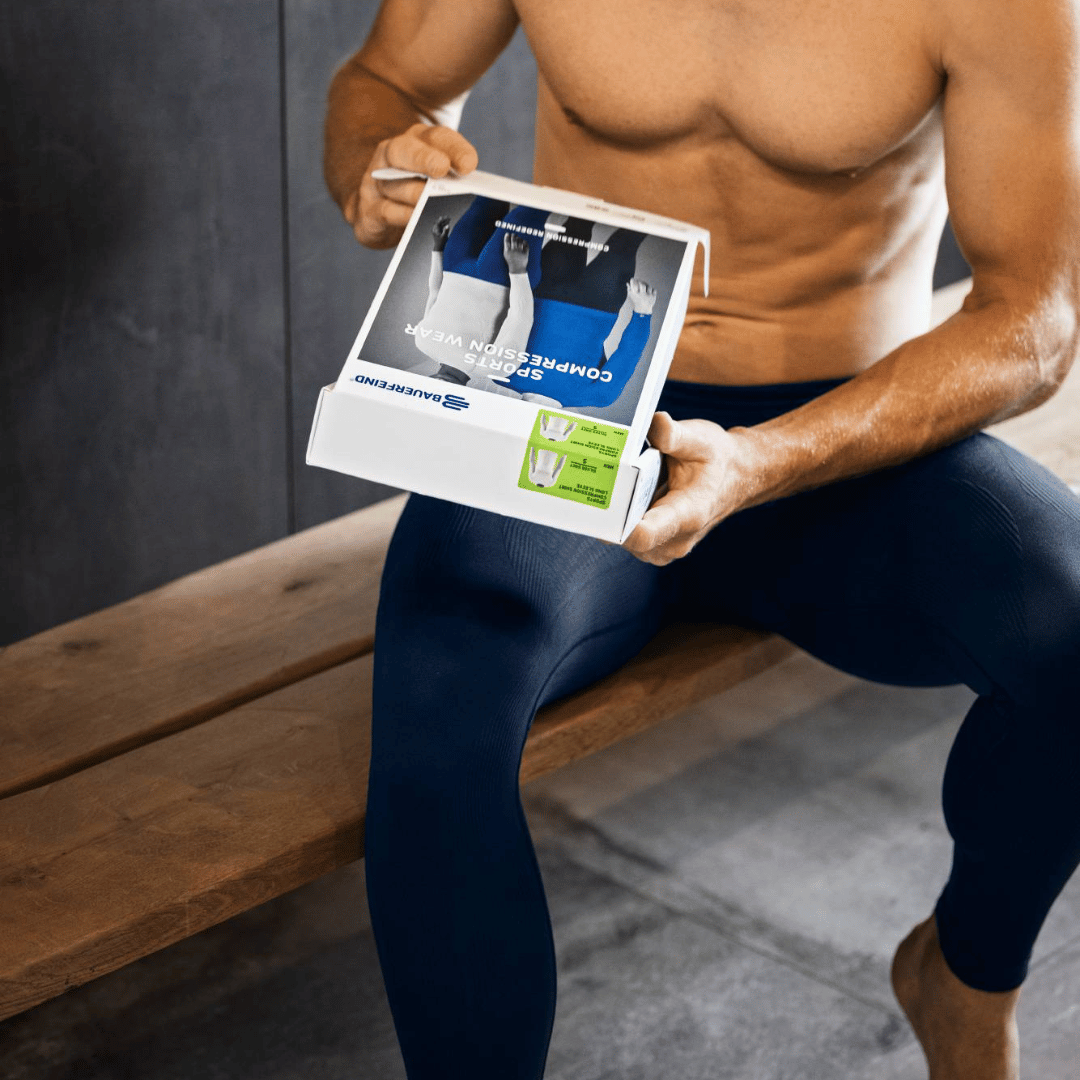 Person holding a product packaging of the bauerfeind compression pants for men , sitting on a wooden bench.