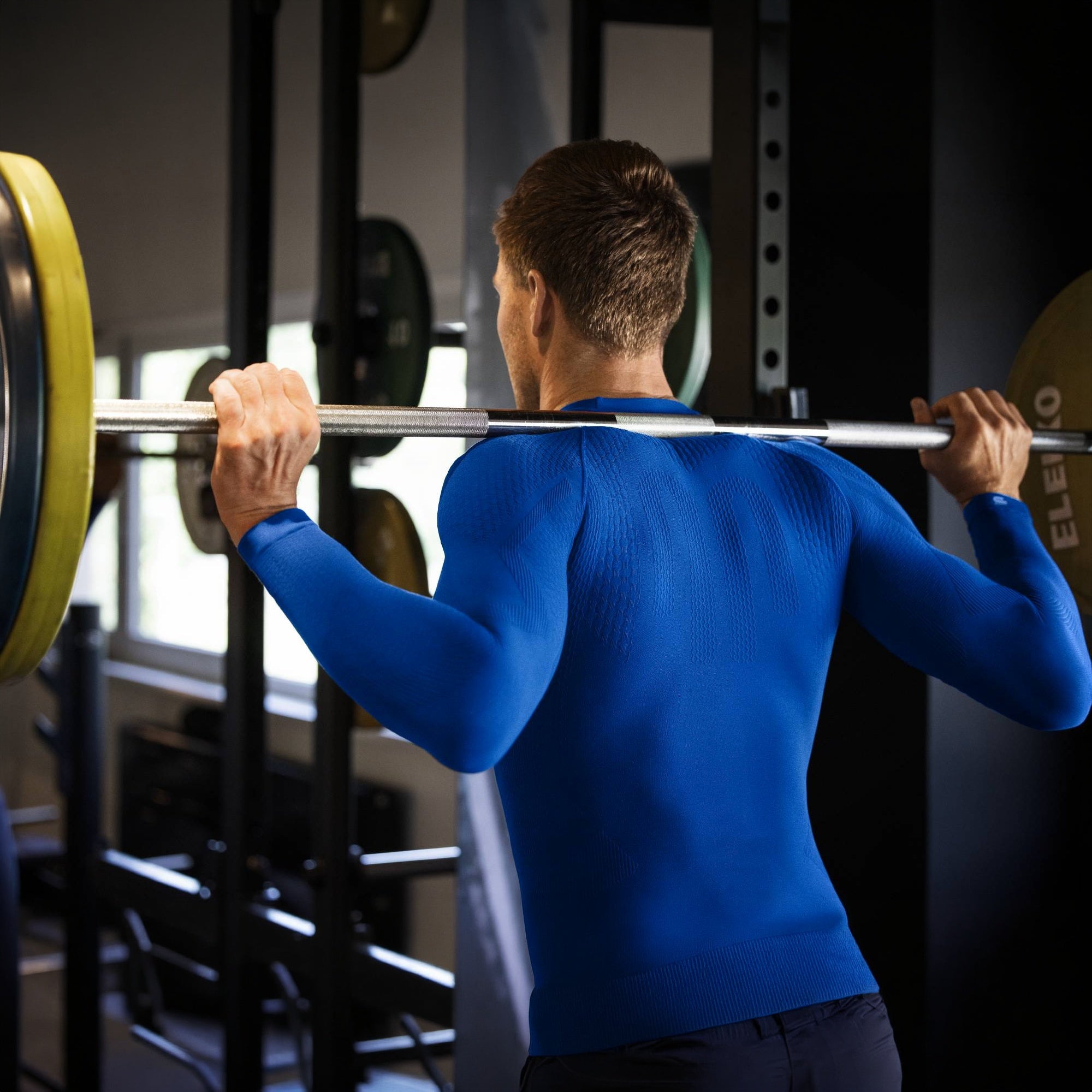 Person lifting a barbell in a gym setting wearing the navy bauerfeind sport men's long sleeve compression shirt