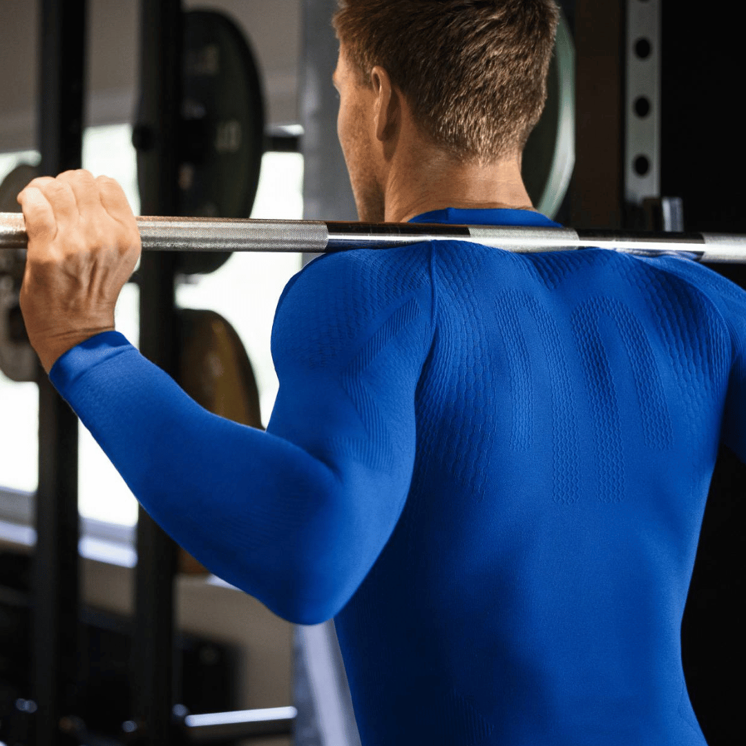 Person in a blue bauerfeind compression shirt long sleeve lifting a barbell in a gym setting
