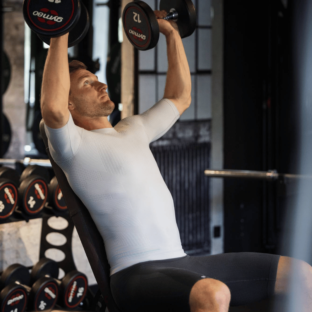 Man wearing bauerfeind's silver sport compression short sleeve shirt and navy sport compression shorts exercising with weights in a gym setting.