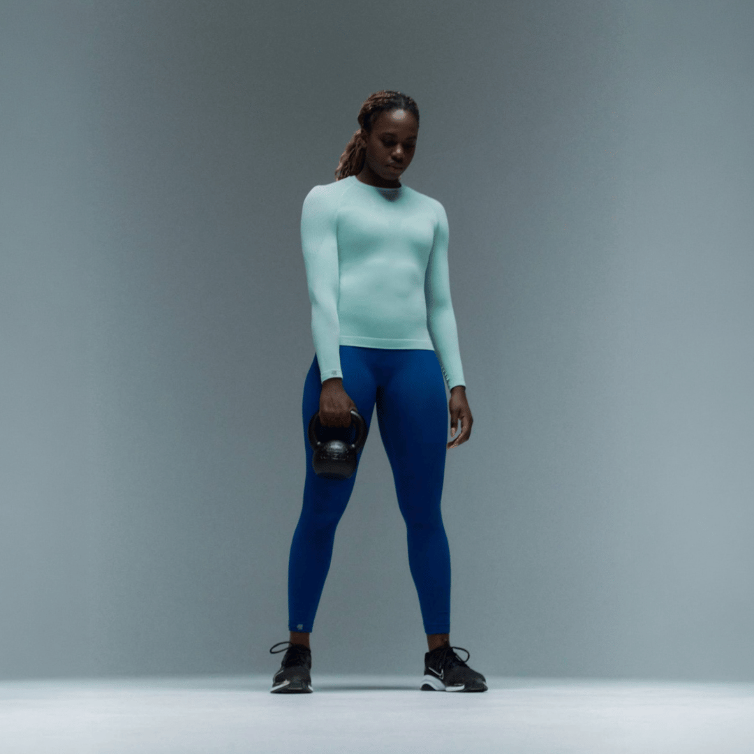 Person wearing a light blue Bauerfeind Women's sport compression long-sleeve top and blue Bauerfeind Women's sport compression leggings holding a black kettlebell against a gray background