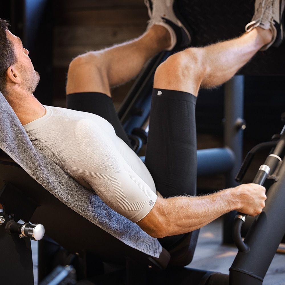 Person in athletic Bauerfeind Grey men's sport compression shorts and in light grey silver Bauerfeind men's sport compression short sleeve shirt, using a leg press machine in a gym setting