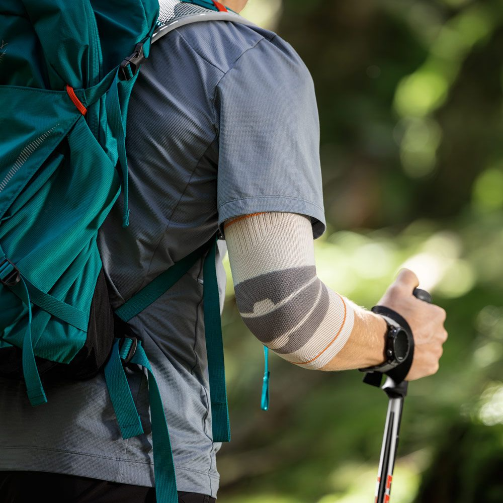 A close-up of a hiker's arm wearing a Bauerfeind Sport Outdoor Elbow compression sleeve, with a teal backpack and hiking pole visible against a blurred forest background.