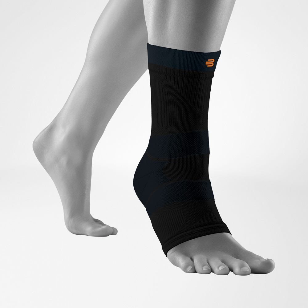 A close-up of a person's ankle wearing a Black Bauerfeind Sports outdoor ankle compression sleeve with a light gray background.