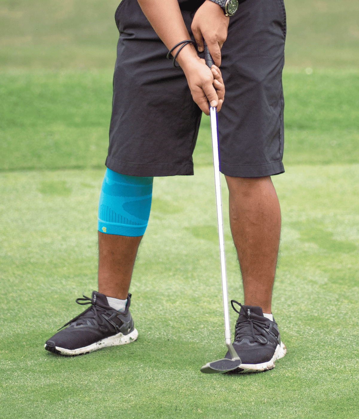 A person on a golf course wearing a bauerfeind compression knee sleeve 20-30 mmhg on one leg, black athletic shorts, and black golf shoes, holding a golf club.