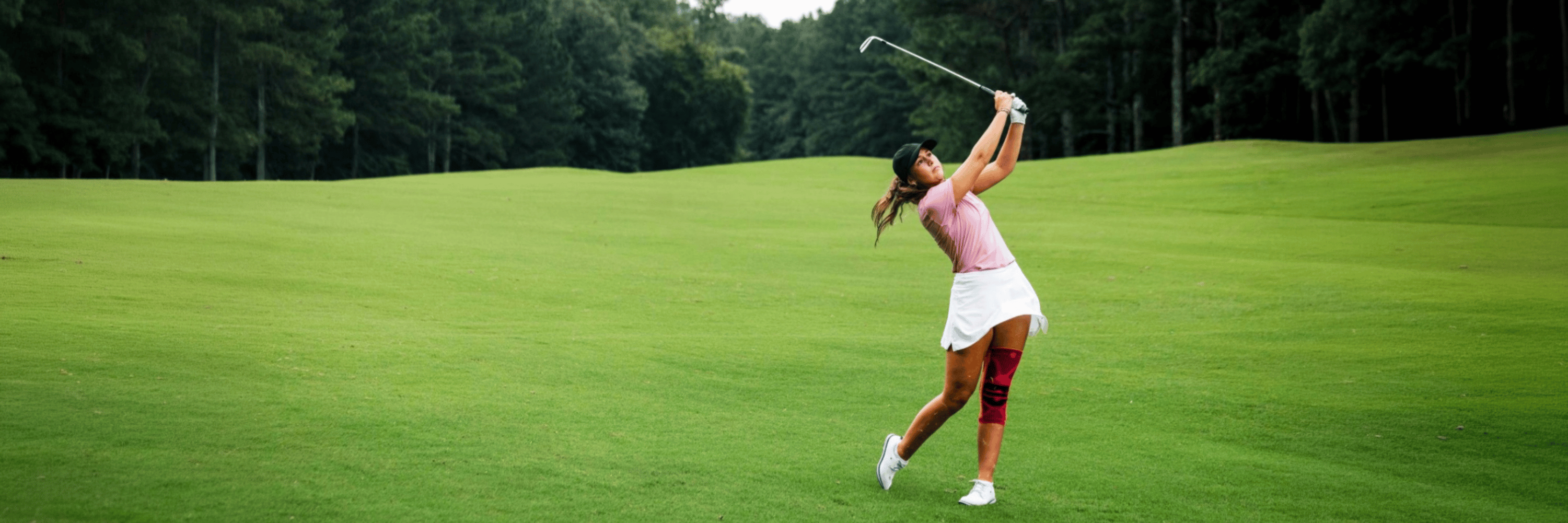 Woman playing golf on a green course with trees in the background wearing a bauerfeind's Pink sports knee support