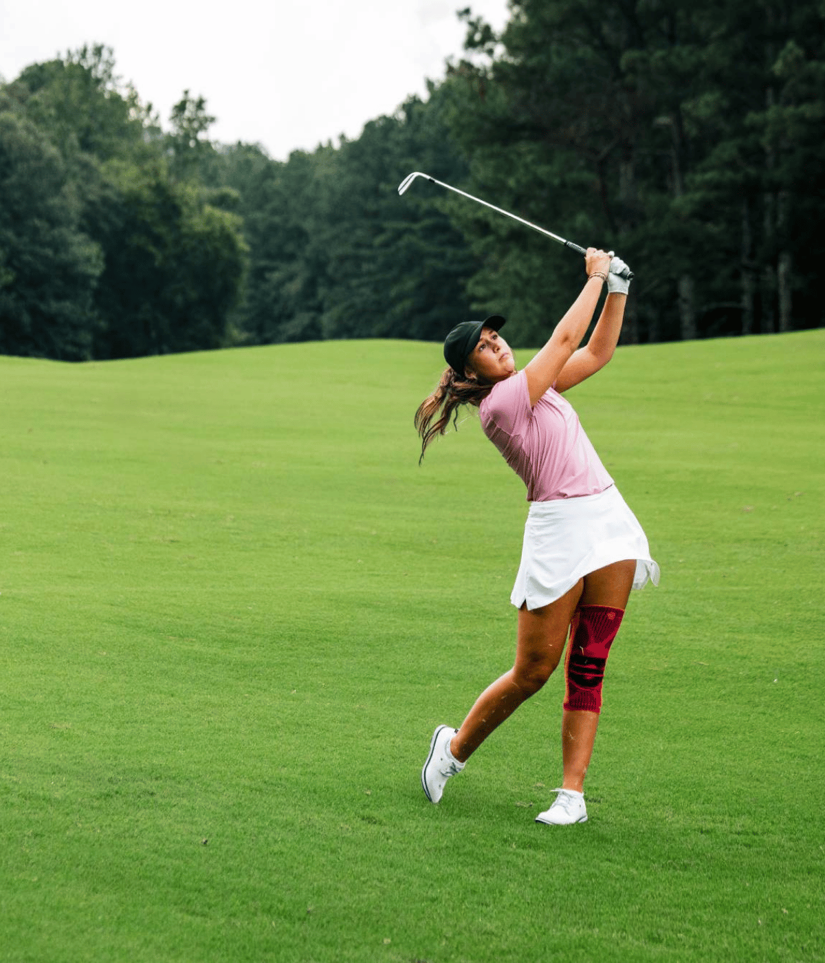 Woman playing golf on a green course with trees in the background wearing a bauerfeind's Pink sports knee support