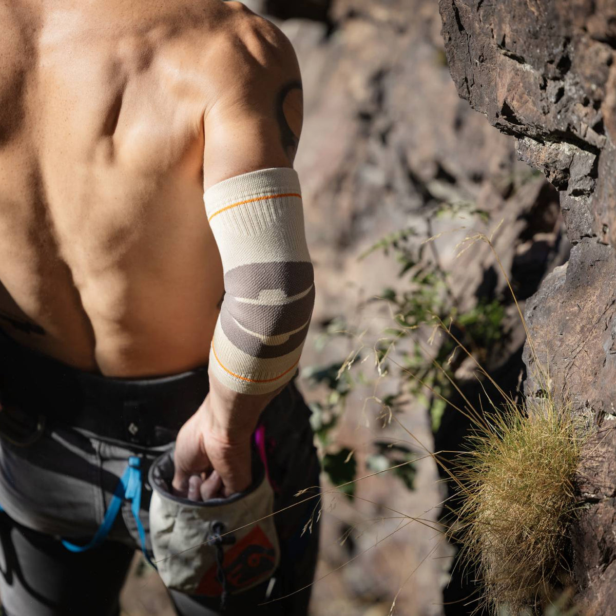 A close-up lifestyle shot of a shirtless male climber from behind, wearing a Bauerfeind Sport Outdoor compression elbow sleeve on his left arm, set against a rocky outdoor cliffside.