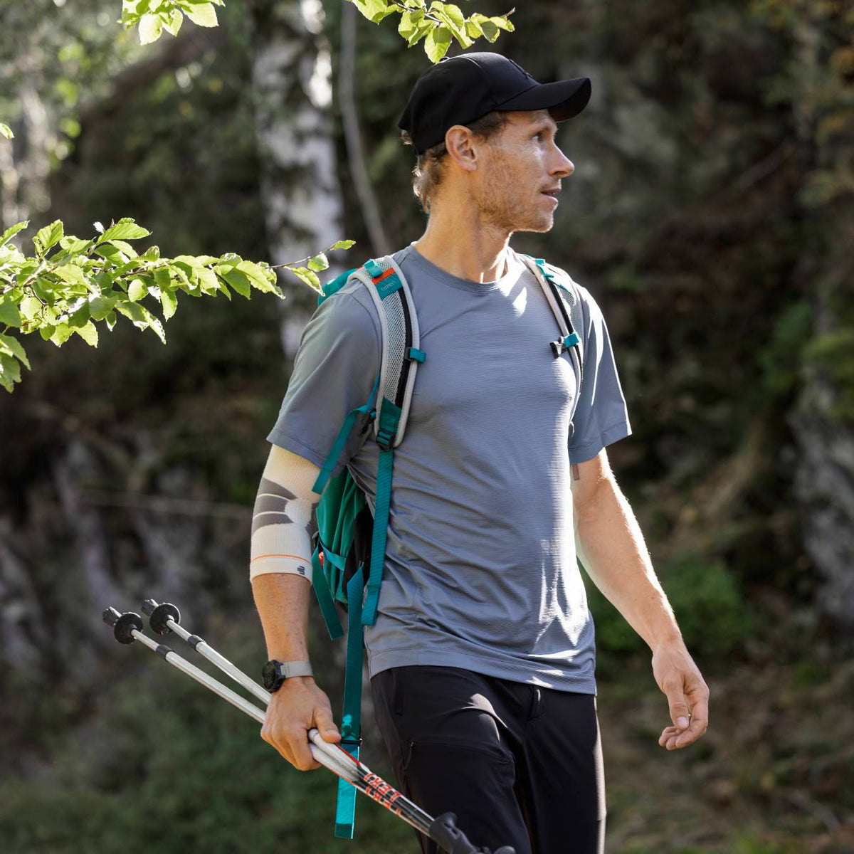 A man hiking in a forest, wearing a Bauerfeind Sport Outdoor Compression Elbow Sleeve with trekking poles.