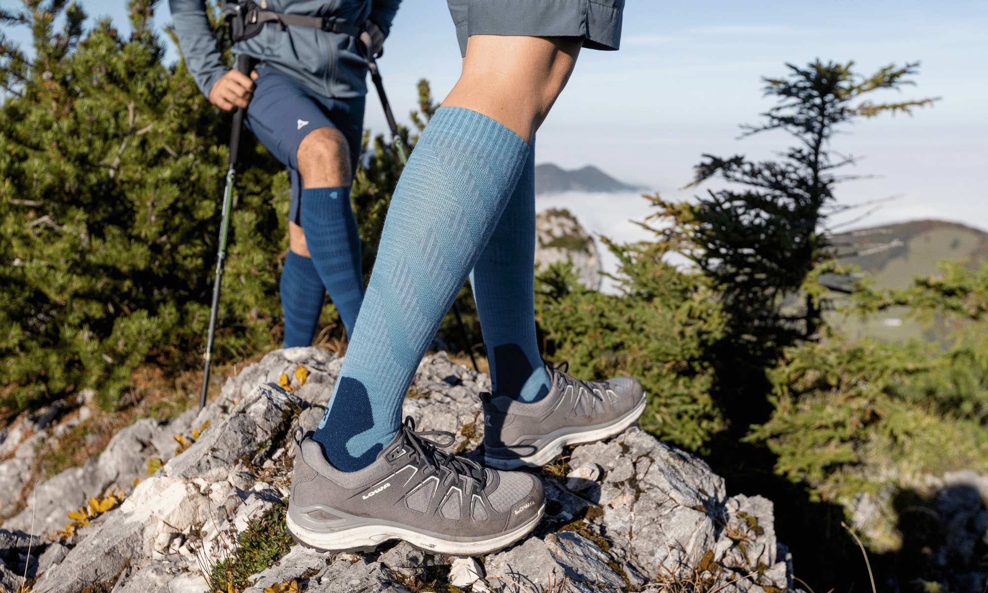 Person hiking on a mountain with Bauerfeind's blue knee-high Outdoor Merino Wool Compression socks and gray shoes.
