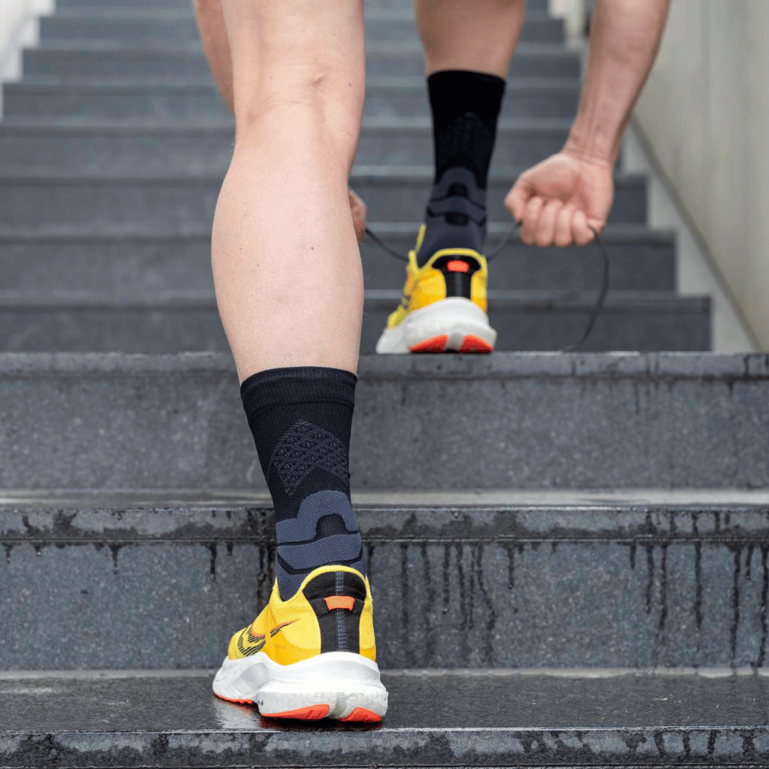 Person wearing Black bauerfeind mid-cut run performance compression socks and yellow running shoes on a concrete surface