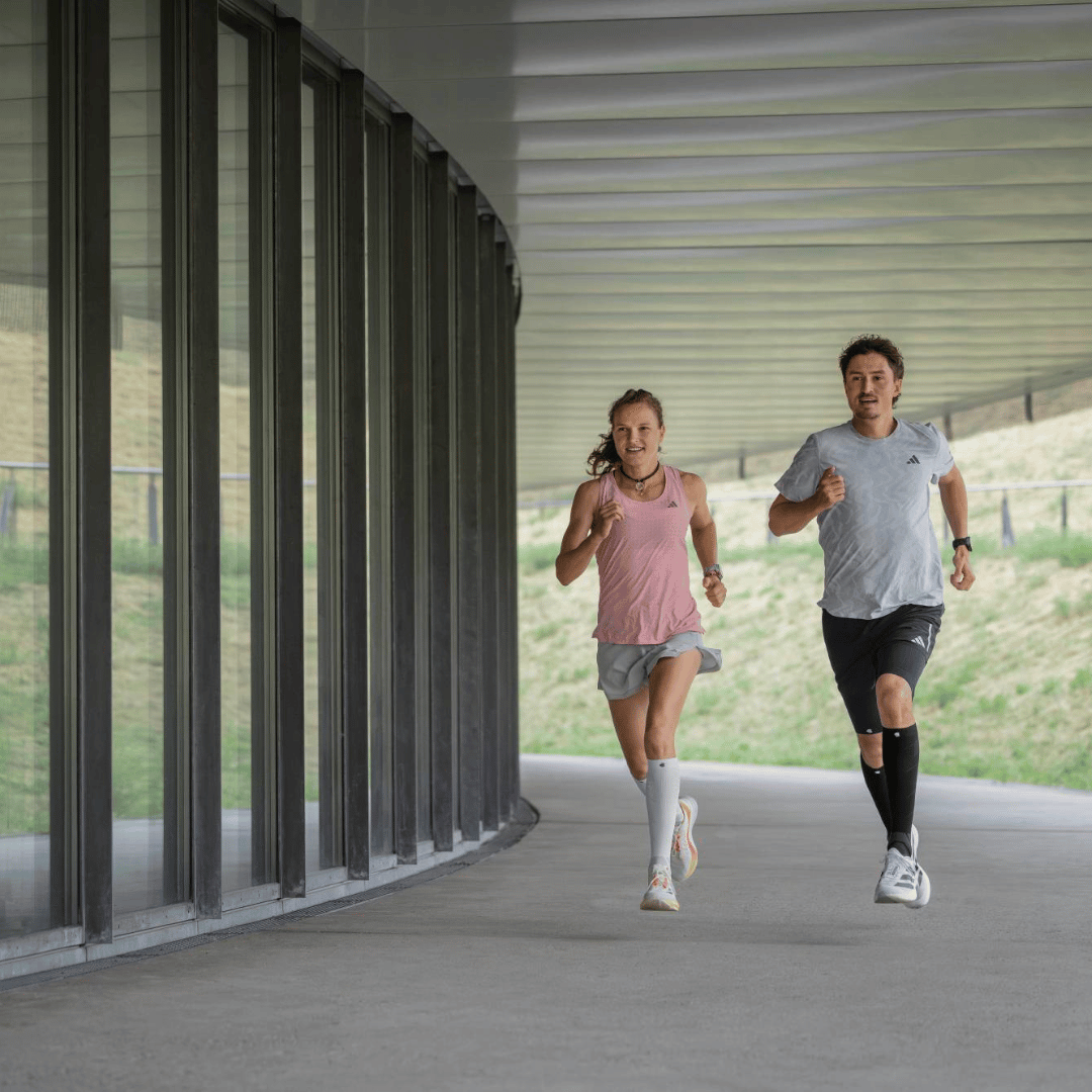 Two people running on a path under a modern architectural structure wearing Bauerfeind Performance Run Compression Socks.