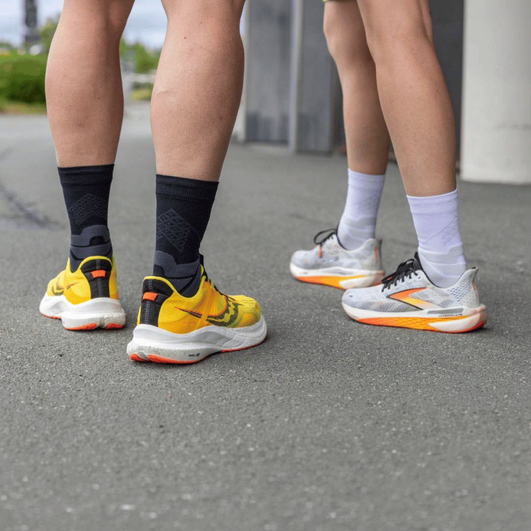 Two people wearing Black and white bauerfeind mid-cut run performance compression socks and different colour  running shoes on a concrete surface