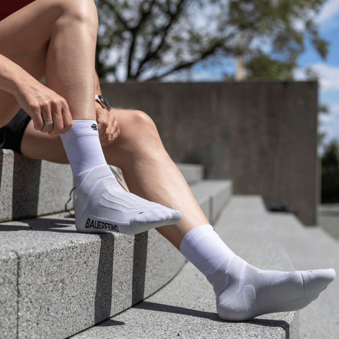 Person wearing white bauerfeind mid-cut run performance compression socks with 'Bauerfeind' branding sitting on concrete steps outdoors.