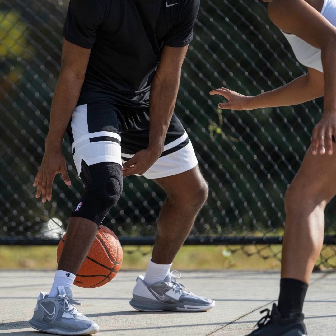 Two people playing basketball on an outdoor court wearing a black Bauerfeind Sports Knee Support NBA with a chain-link fence and trees in the background.