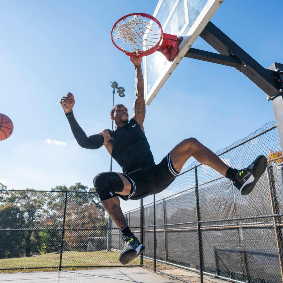 a person playing basketball on an outdoor court wearing a black Bauerfeind Sports Knee Support NBA with a chain-link fence and trees in the background.