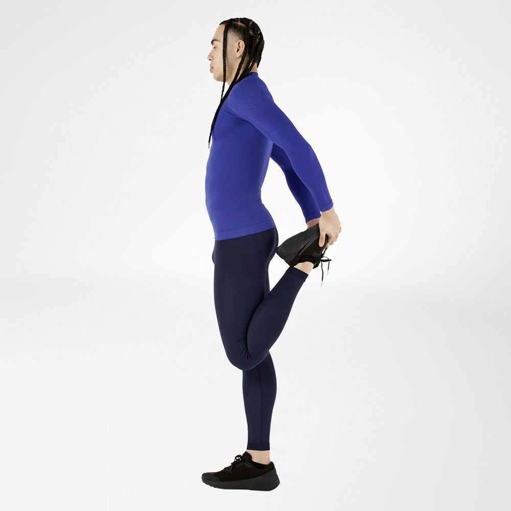 Person wearing a blue long-sleeve bauerfeind compression shirt and navy mens bauerfeind compression leggings, stretching on a white background