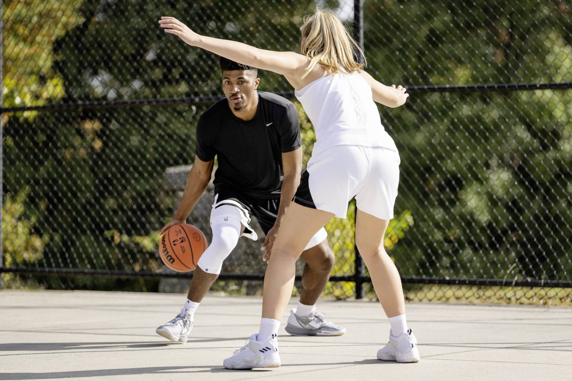 Two people playing basketball on an outdoor court wearing a white bauerfeind Sports Compression Knee Support NBA with a chain-link fence and trees in the background.