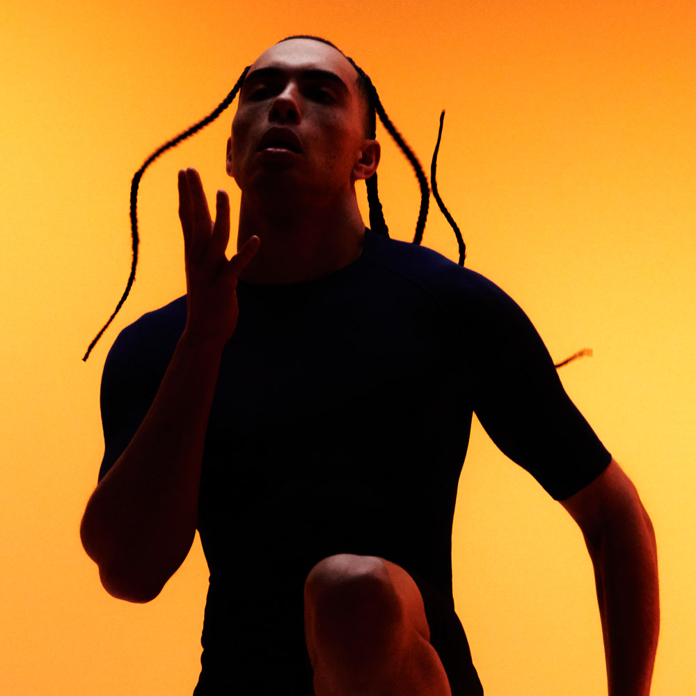 Person with braided hair wearing bauerfeind sports compression wear shorts and short sleeve shirt against an orange background